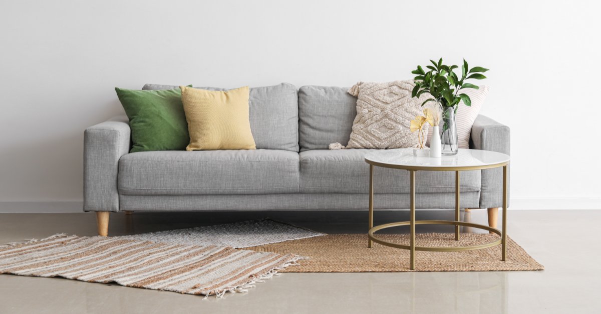 A gray sofa with green and yellow pillows, a white table, plant-filled vases, and layered rugs against a plain wall.