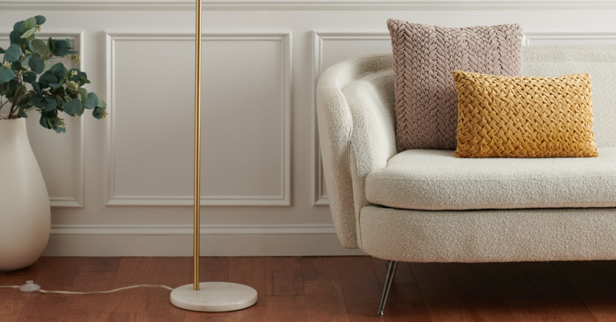 A cream loveseat with woven pillows beside a gold floor lamp and potted plant on a wood floor by paneled walls.