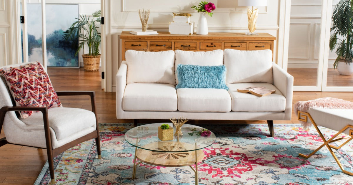 A bright living room with a white sofa, patterned rug, glass coffee table, armchair, plants, and wooden floors.