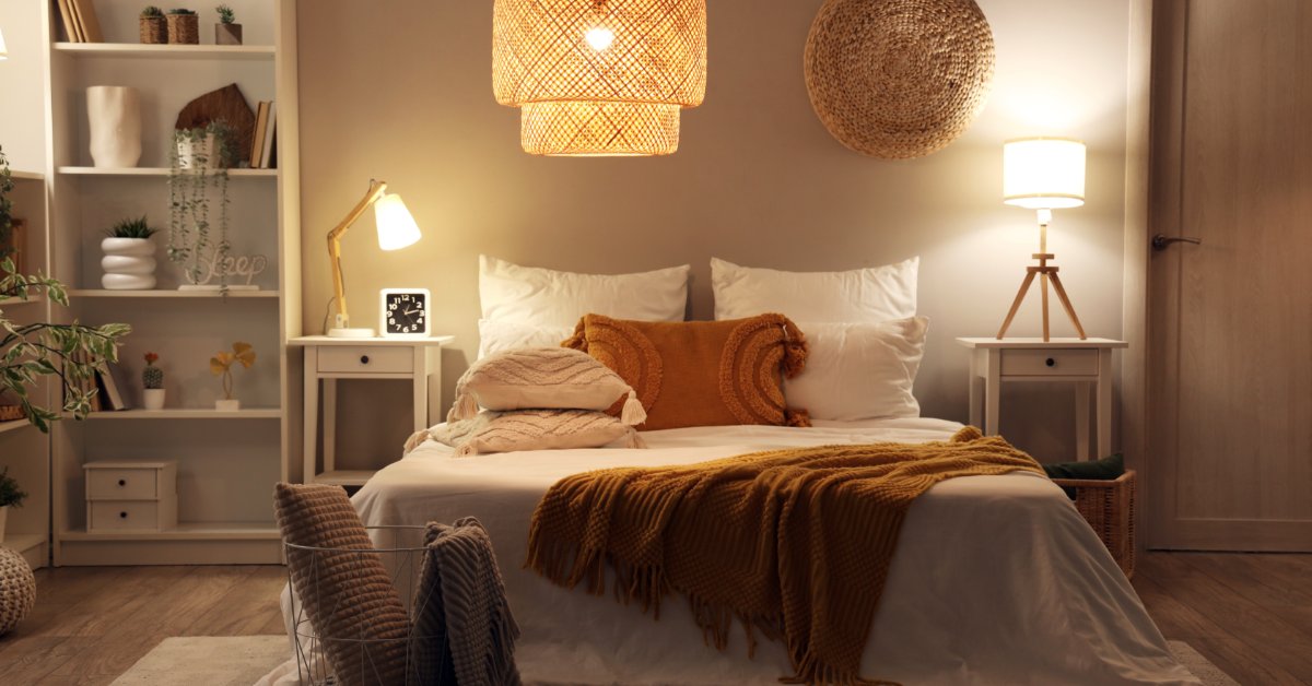 A cozy bedroom with a white bed, orange blanket, woven pendant light, shelves, nightstands, and lit lamps.